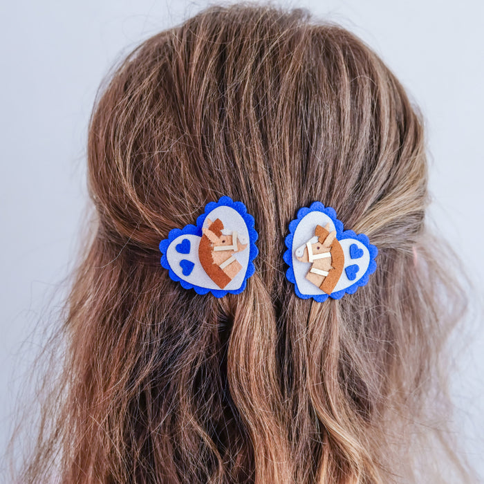 Two blue and white heart-shaped hair clips with small beige ribbon accents, shown pinned to the back of a person's brown hair against a light gray background.