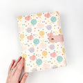 Hand holding the Heart Yarn Ball Project Journal Planner showing the cream faux leather cover printed with colorful heart-shaped yarn balls, round yarn skeins, and stars in pink, teal, purple, and gold with a pink snap closure