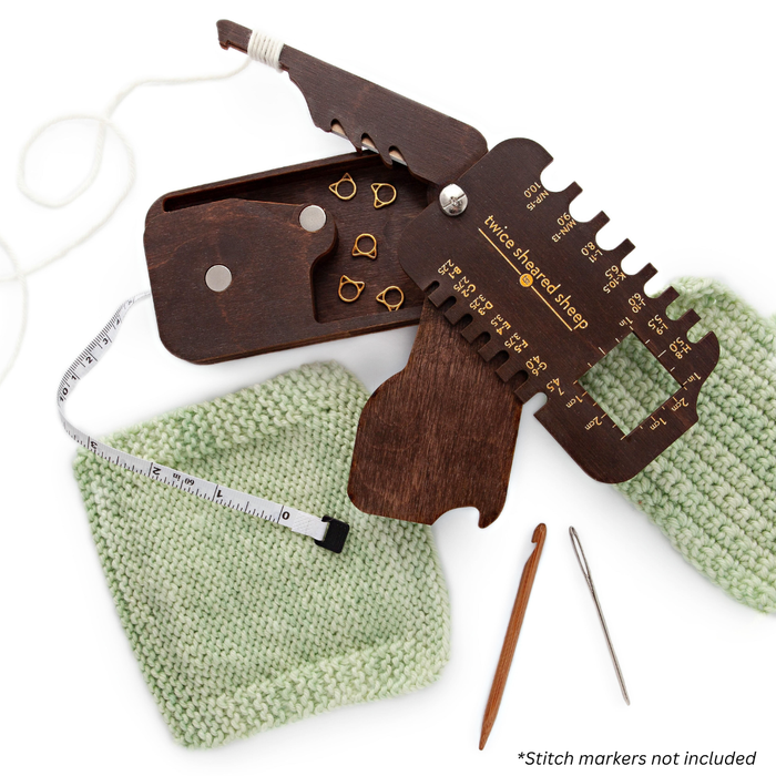 Stitch Scout Multi-Tool opened on a green knitted swatch showing needle gauge, storage box with stitch markers, measuring tape, cable needle, and darning needle