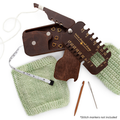 Stitch Scout Multi-Tool opened on a green knitted swatch showing needle gauge, storage box with stitch markers, measuring tape, cable needle, and darning needle