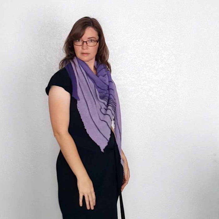 Front view of model wearing the Primus shawl in purple and lavender draped loosely around her neck over a black dress, showing the dark striped texture