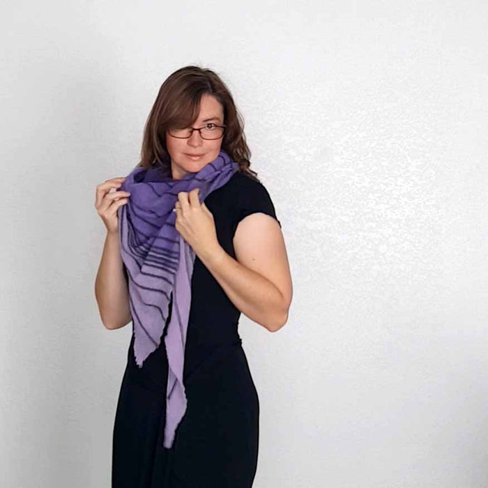 Model adjusting the Primus shawl in purple and lavender wrapped around her neck, holding the fabric with one hand over a black top