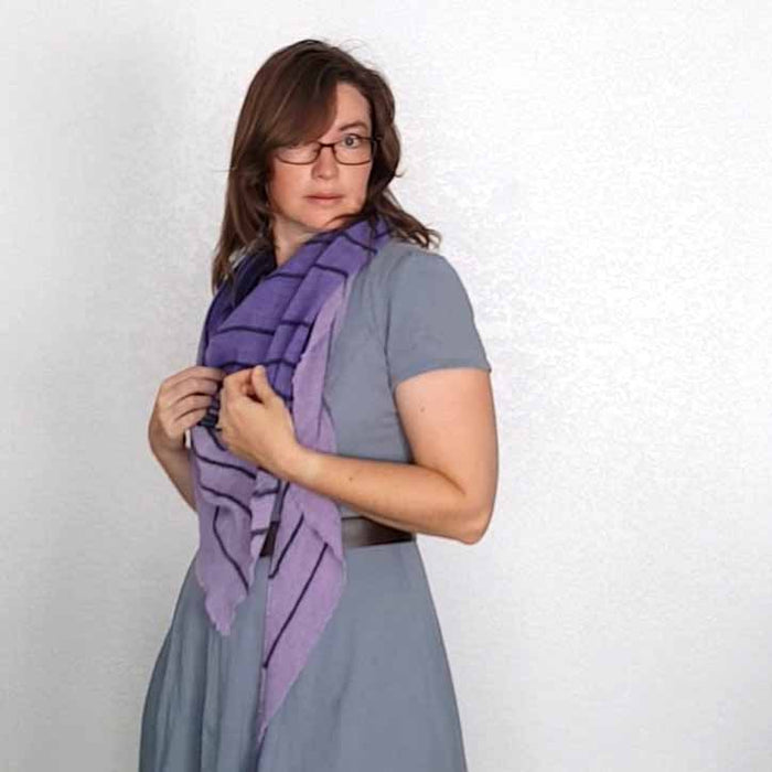 Side view of model wearing the Primus shawl in purple and lavender around her neck over a gray-blue dress, holding the ends with both hands