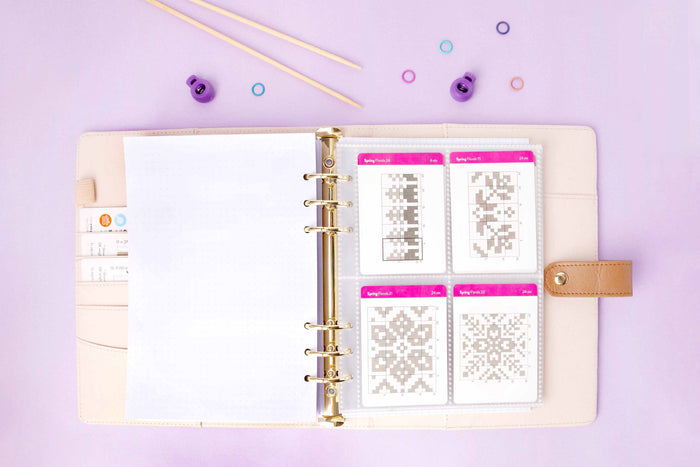 Project journal open showing four colorwork chart cards stored in a clear four-pocket card protector sleeve on lavender background with knitting accessories