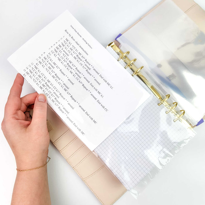 Hand holding a printed knitting pattern page inside a clear protector sleeve in the project journal showing row-by-row colorwork instructions