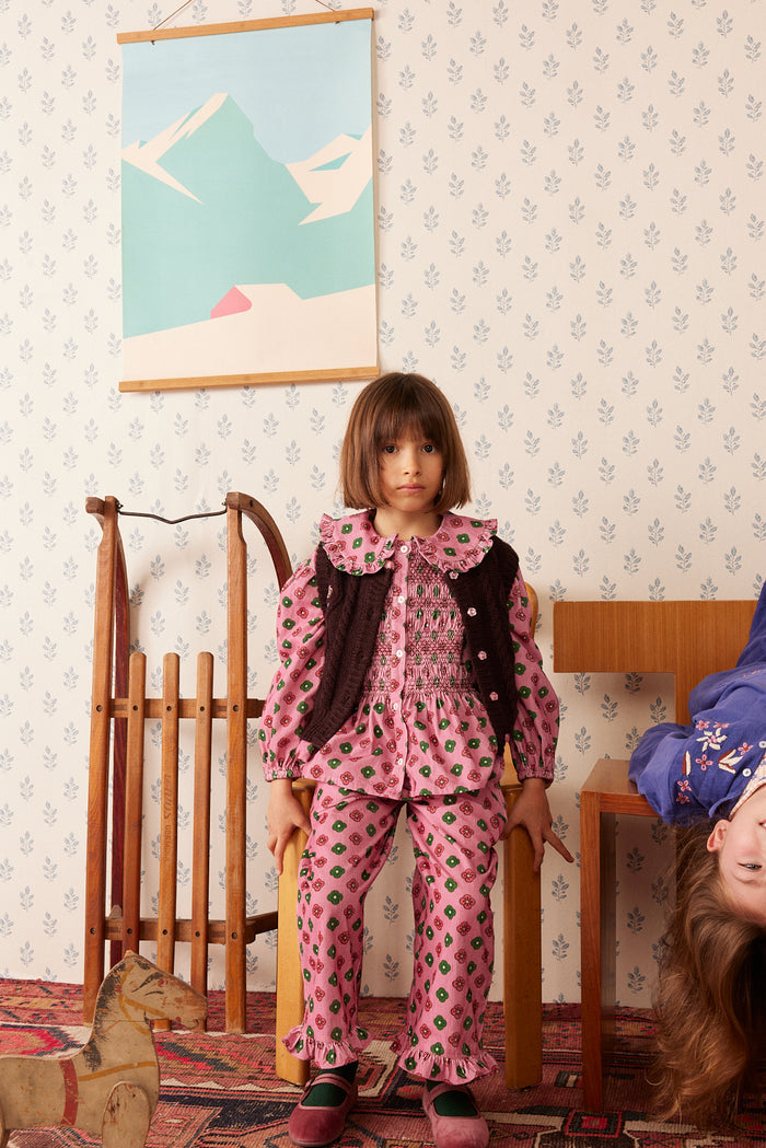 A young girl stands in a vintage-style room wearing a pink patterned pajama set with a dark brown vest. The room features patterned wallpaper, a wooden sled, and a framed landscape print on the wall.