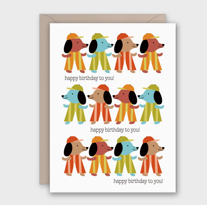 A birthday greeting card with a kraft envelope behind it. The card features a grid of cartoon dogs wearing colorful striped outfits and the text 'happy birthday to you!' printed below each dog.