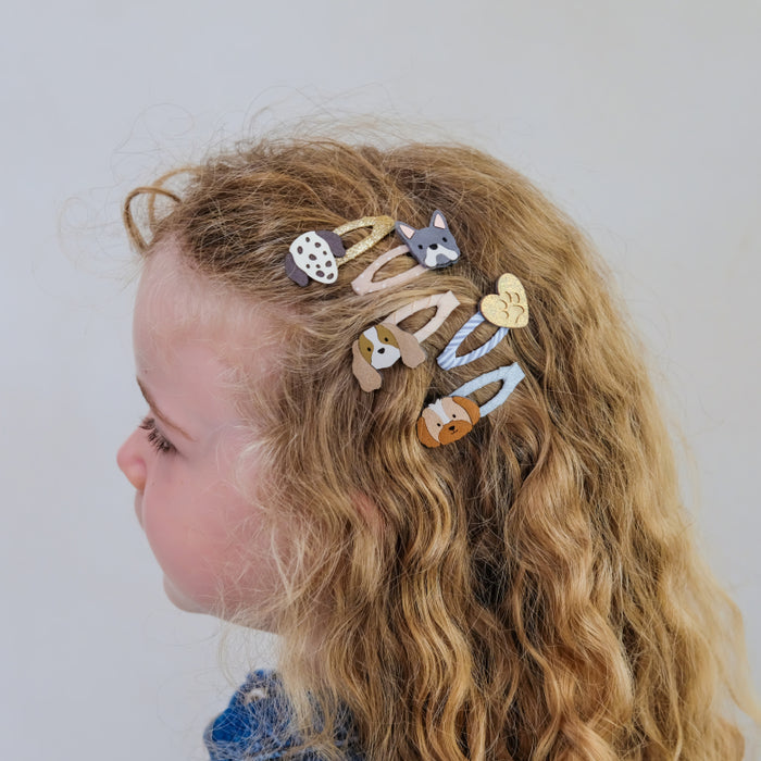 A side-profile view of a young girl with long, wavy blonde hair wearing six decorative hair clips shaped like animals and objects, including a fox, a duck, a heart, and a polka-dot pattern.