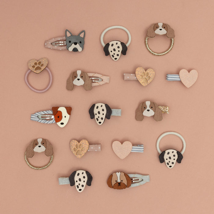 A set of dog-themed hair clips arranged on a solid dusty pink background. The clips feature various dog face designs, heart-shaped charms, and ring-style accents in brown, beige, black, white, and light pink.