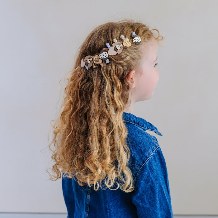 A young girl with long, curly blonde hair is shown from the side profile, wearing a decorative hair clip adorned with wooden hearts and small bows in her hair. She is wearing a blue denim shirt against a plain light gray background.