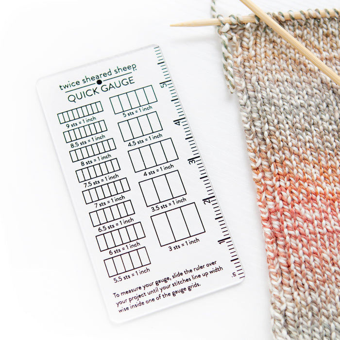 Quick Gauge ruler placed beside multicolor knitting swatch on bamboo needles showing gauge measurement in use