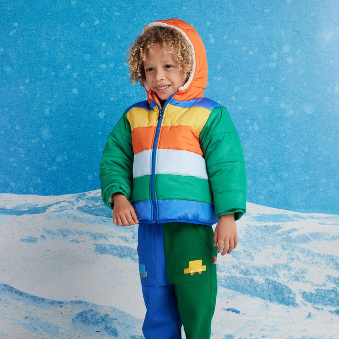 Child wearing a colorful striped jacket and green pants with a yellow car design, standing against a blue snowy background.
