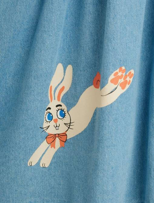 A close-up of a light blue textured fabric, likely denim, featuring a cartoon rabbit graphic. The rabbit is cream-colored with blue eyes, an orange bow tie, and orange paw pads, set against a background of blue fabric folds.