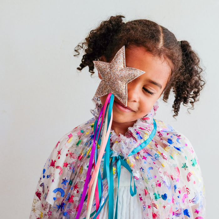 A young girl with curly brown hair in pigtails wears a sparkly silver glitter star wand over her eye. The wand has long, flowing pastel ribbons in pink, blue, and purple.
