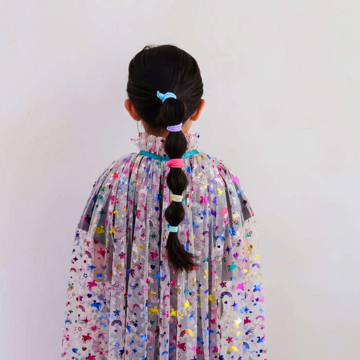 A child viewed from behind wearing a sheer, pleated cape or dress covered in colorful star and character prints, with dark hair styled in a braid and secured with pastel hair ties against a plain light background.