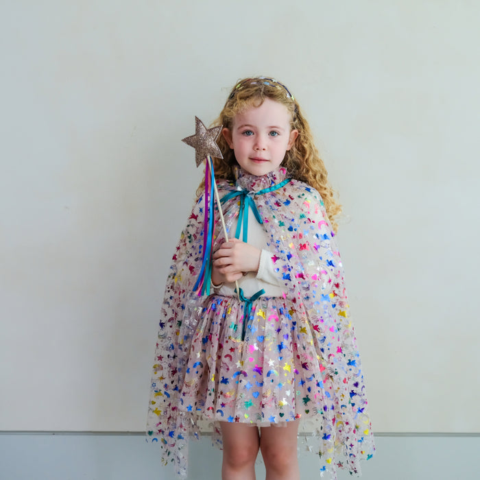 A young girl with curly blonde hair stands against a plain light gray wall wearing a sparkly multicolor fairy cape dress over a white long-sleeve top. The dress is covered in holographic unicorn and rainbow confetti, with a teal bow at the collar and waist.