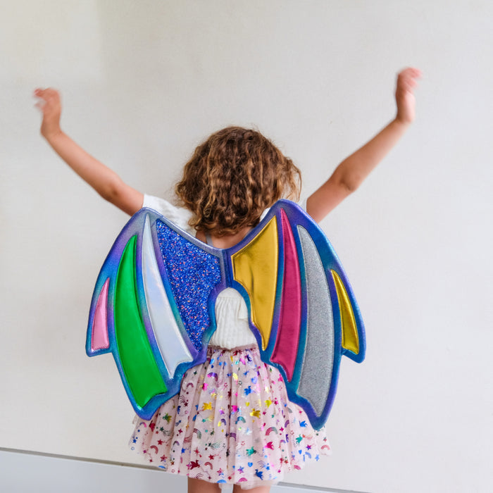 A child seen from behind wearing a colorful angel wings costume with glittery blue, green, pink, yellow, and silver wings, paired with a white t-shirt and a pink star-patterned tutu skirt, with arms raised against a plain white wall.