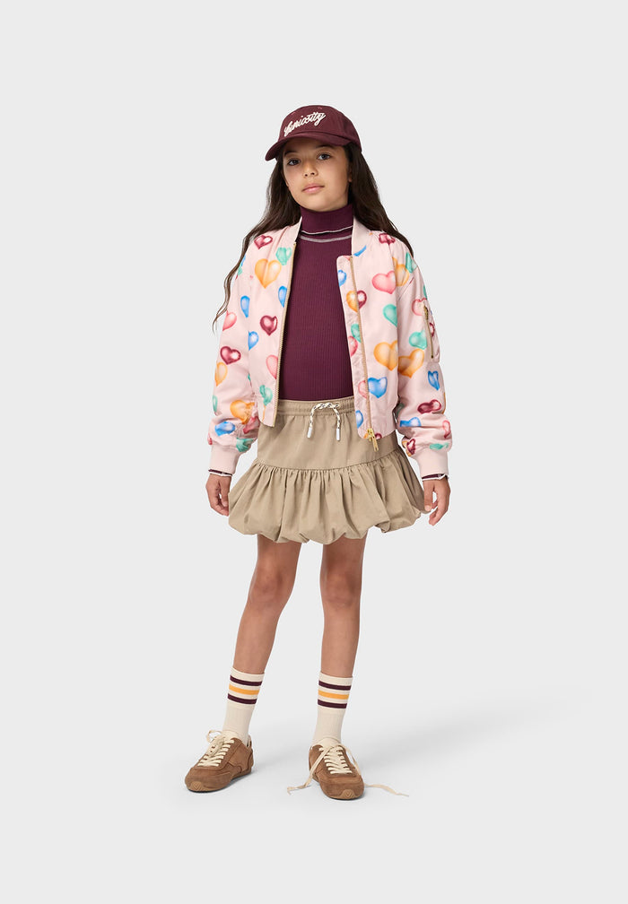 Young girl wearing a colorful jacket, purple shirt, beige skirt, and maroon cap on a white background