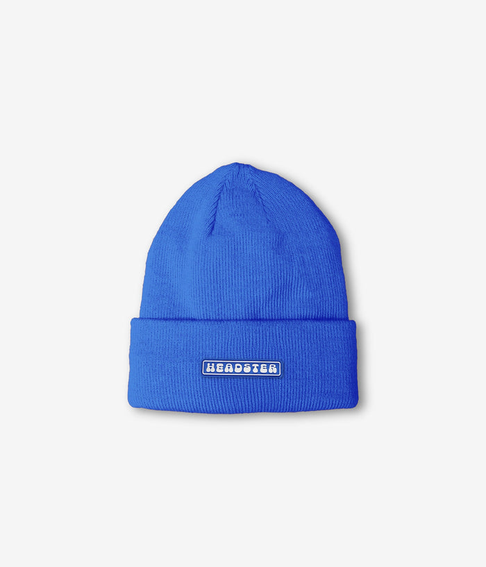 A bright blue ribbed knit beanie hat with a folded cuff, featuring a small rectangular white patch with the word 'HEADSTER' in blue text, shown on a light gray background.