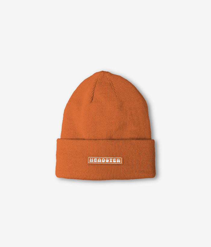 An orange ribbed knit beanie hat with a folded cuff, featuring a small rectangular white patch on the front with the word 'HEADSTER' in white text.