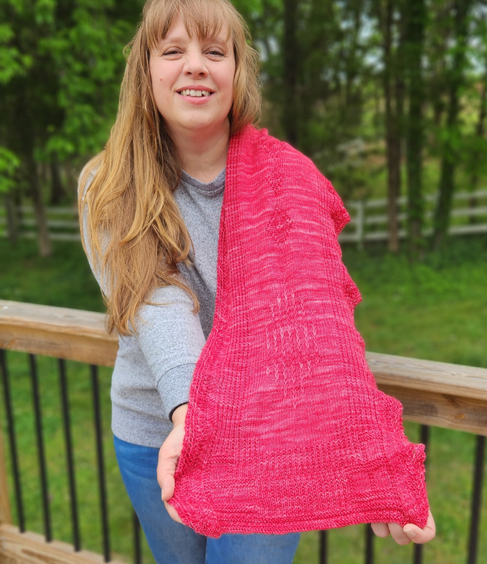 Woman holding the Revenge in Lido Wrap in coral pink lace knit fabric up to show the drape and openwork stitch pattern