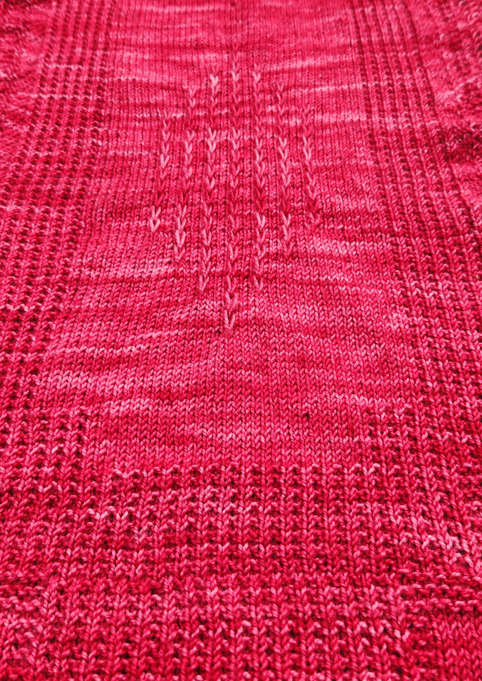Woman holding the Revenge in Lido Wrap up flat showing the full rectangular shape and allover lace stitch pattern in coral pink fingering weight yarn