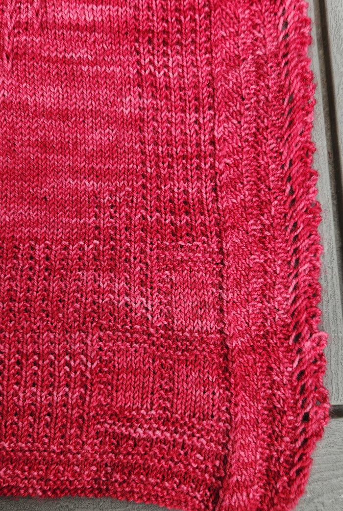 Woman holding the Revenge in Lido Wrap in coral pink up near her face showing the delicate lace openwork stitch pattern