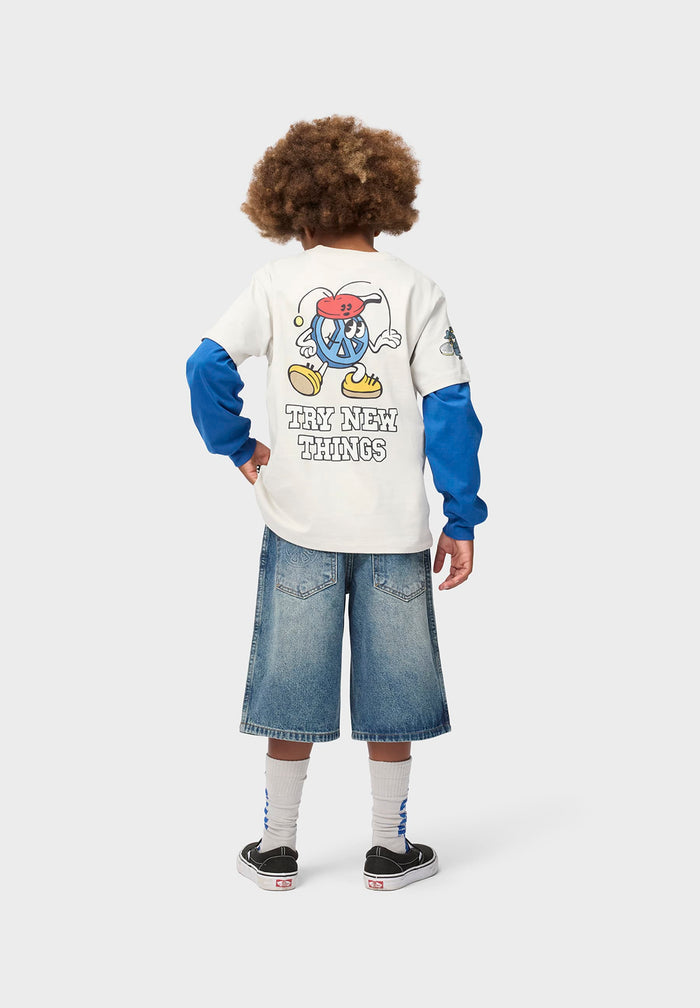 Child wearing a white long-sleeve shirt with a cartoon character and text, blue sleeves, and denim shorts on a white background.