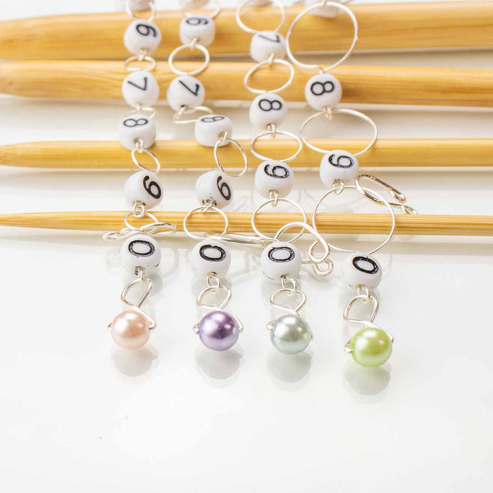 Four numbered bead chain row counters with pastel pearl charm ends in pink, purple, silver, and green hanging from bamboo knitting needles