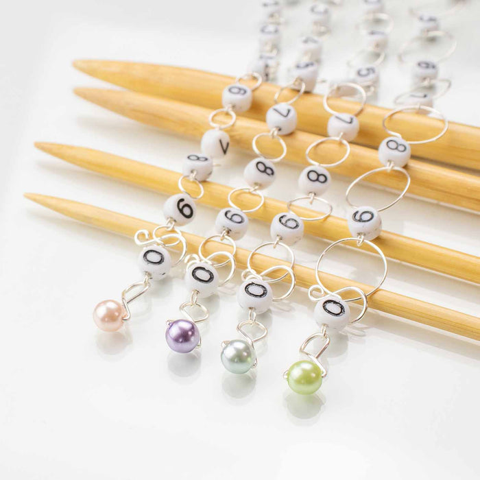 Close-up of four row counter chain ends showing pastel pearl beads in pink, purple, silver, and green with numbered beads on bamboo needles