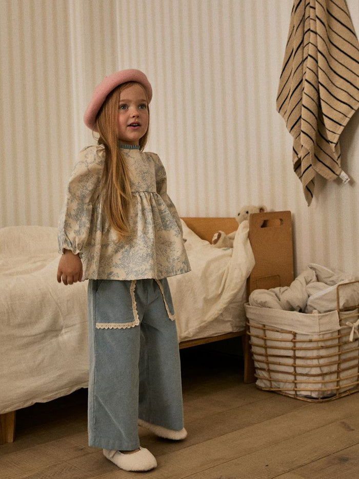 Young girl in a patterned top and blue pants standing in a bedroom.