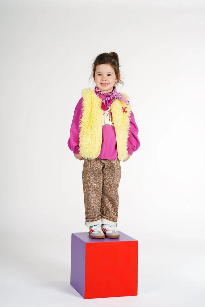 child standing on purple and red box wearing yellow faux fur vest, fuchshia sweatshirt and bandana, with leopard printed trousers and red and white sneakers.