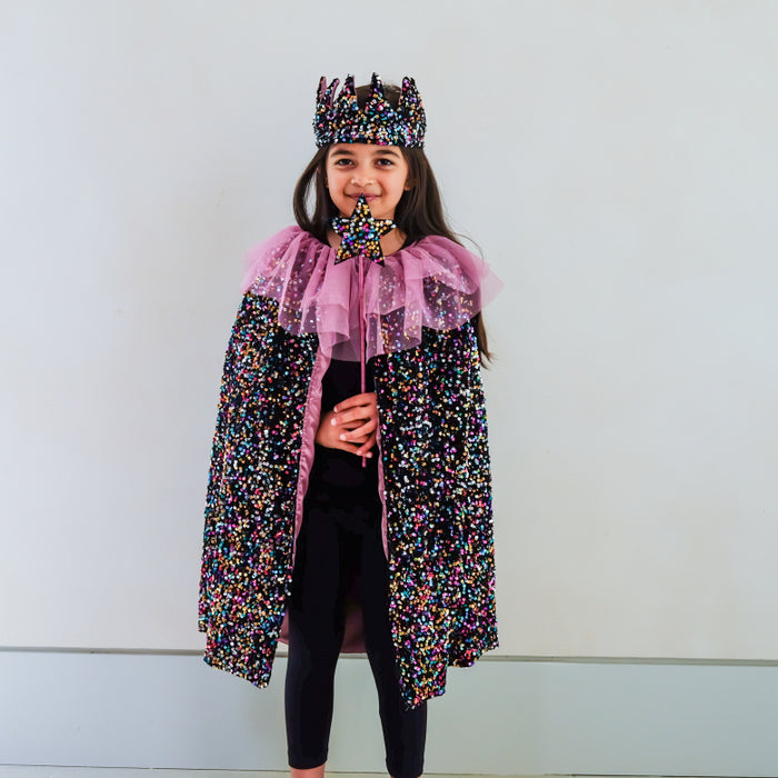 A young girl wearing a black sequined royal cape with a mauve lining, a matching black sequined crown, and a sequin wand, standing against a plain light gray wall.