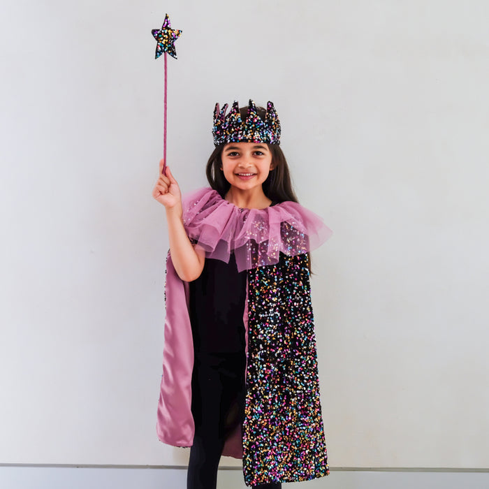 A young girl wearing a purple and black sequined fairy/princess costume with a matching crown and holding a star wand, standing against a plain white wall.