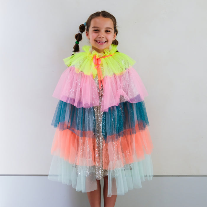 A young girl with braided hair wearing a colorful, tiered tulle cape with neon yellow, pink, teal, orange, and light blue layers over a sparkly silver dress, standing against a plain white wall.