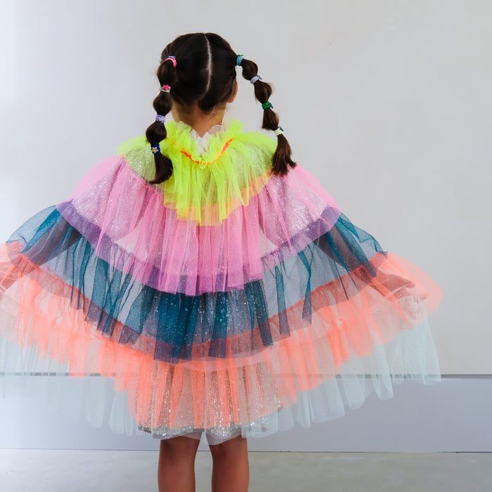 A child viewed from behind wearing a voluminous layered tulle cape with bright neon pink, lime green, blue, peach, and white sheer layers against a plain light gray wall.
