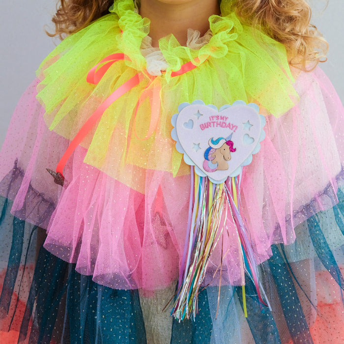 A close-up of a child wearing a colorful tulle cape with layered neon yellow, pink, and blue sparkly layers, plus a heart-shaped 'It's My Birthday!' party accessory with multicolored streamers.