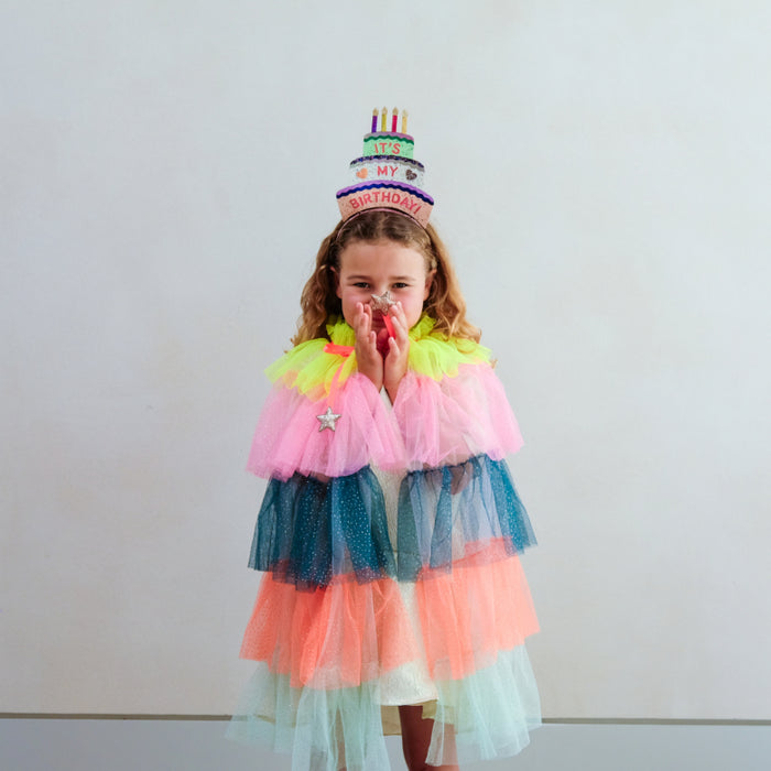 A young girl wearing a colorful tiered tulle cape with neon pink, blue, orange, and white layers, along with a matching birthday hat shaped like a cake, standing against a plain light gray wall.