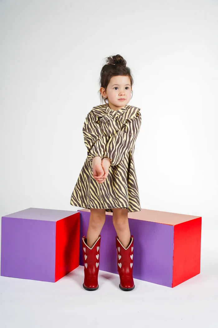 child standing in front of red and purple boxes wearing yellow and brown zebra stripe dress with peter pan collar and bow with red cowboy boots on white background.
