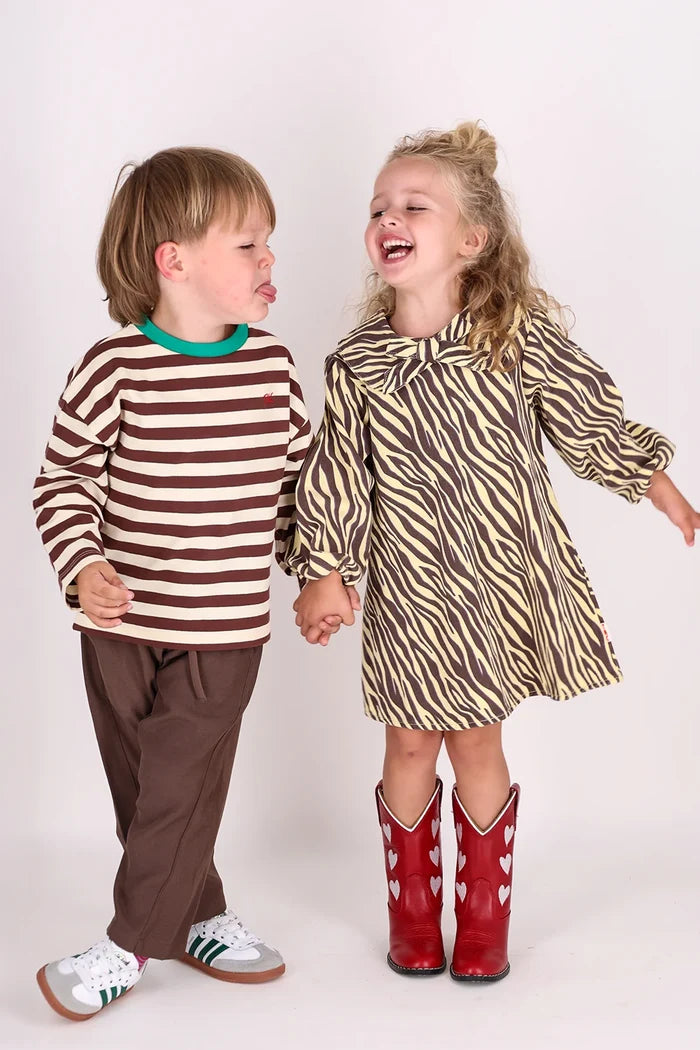 child standing wearing yellow and brown zebra stripe dress with peter pan collar and bow with red cowboy boots on white background.
