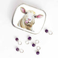 Small hinge-style Sassy Sheep storage tin open beside purple amethyst bead infinity-loop snag-free stitch markers on white background