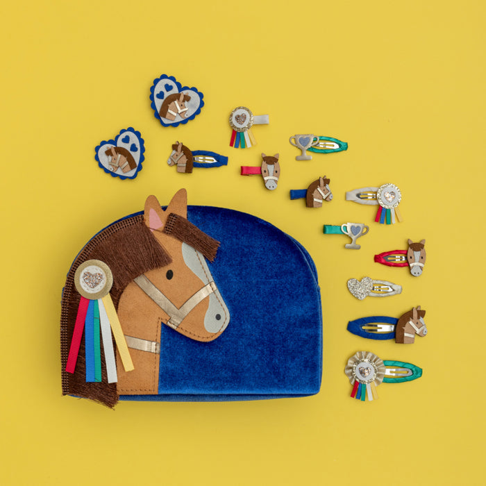 A flat-lay product shot of a horse-themed hair accessory set on a solid yellow background. The set includes a blue velvet pouch shaped like a horse's head with brown and tan leather details, alongside multiple matching hair clips and pins in horse, ribbon, and trophy designs.