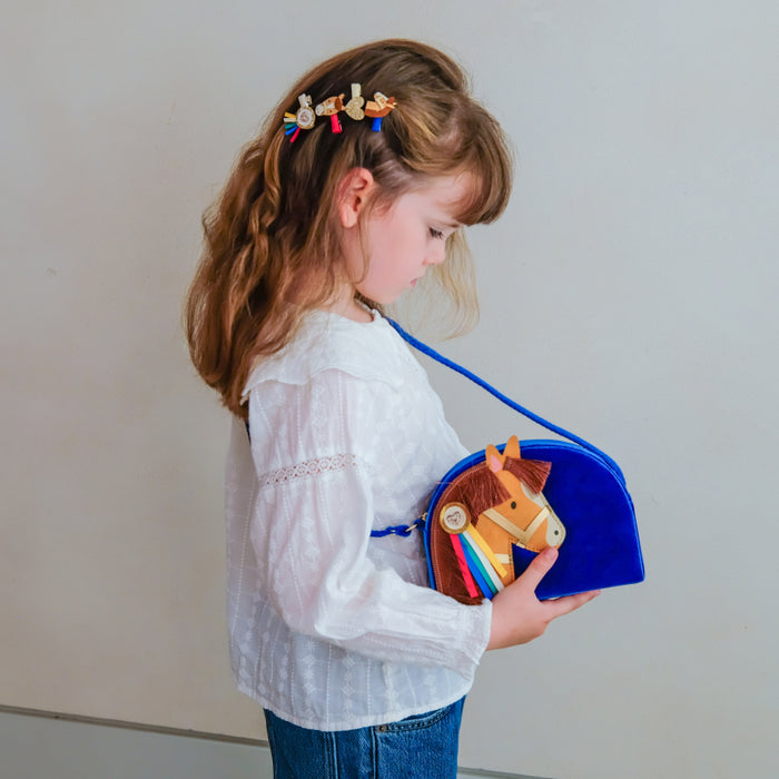 A young girl with light brown hair wearing a white long-sleeve blouse and blue jeans is shown in profile, holding a blue felt horse-shaped bag with a wooden rocking-horse face and colorful tassels. She has small decorative hair clips in her hair.