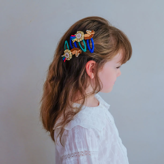 A young girl with light brown hair is shown in profile wearing three colorful hair clips with gold-tone floral details. The hair clips feature blue, green, red, and gold elastic bands.