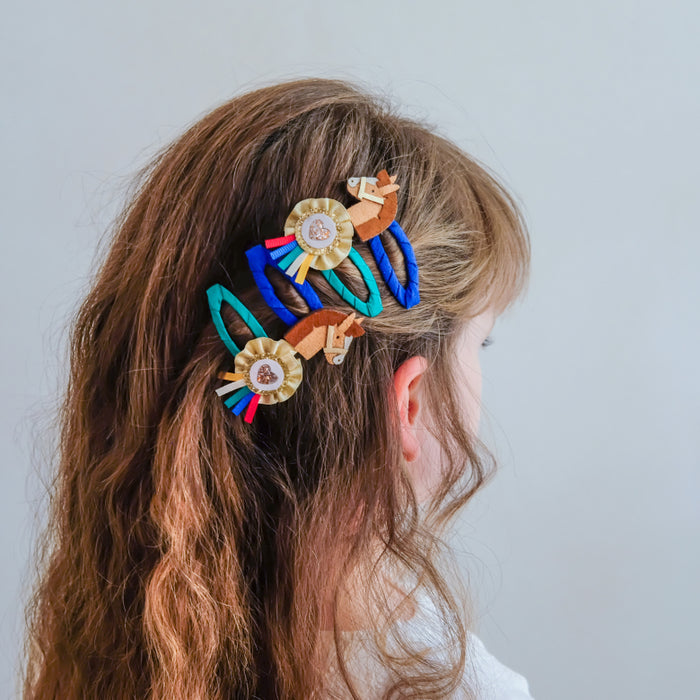 A side profile of a young girl with long wavy light brown hair, wearing a set of colorful decorative hair clips with horse motifs and gold medallion details, against a plain light gray background.