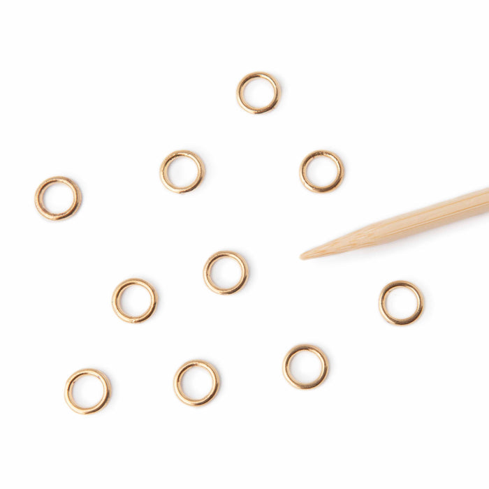 Ten simple gold ring stitch markers scattered on a white background next to a bamboo knitting needle tip showing the small closed ring size