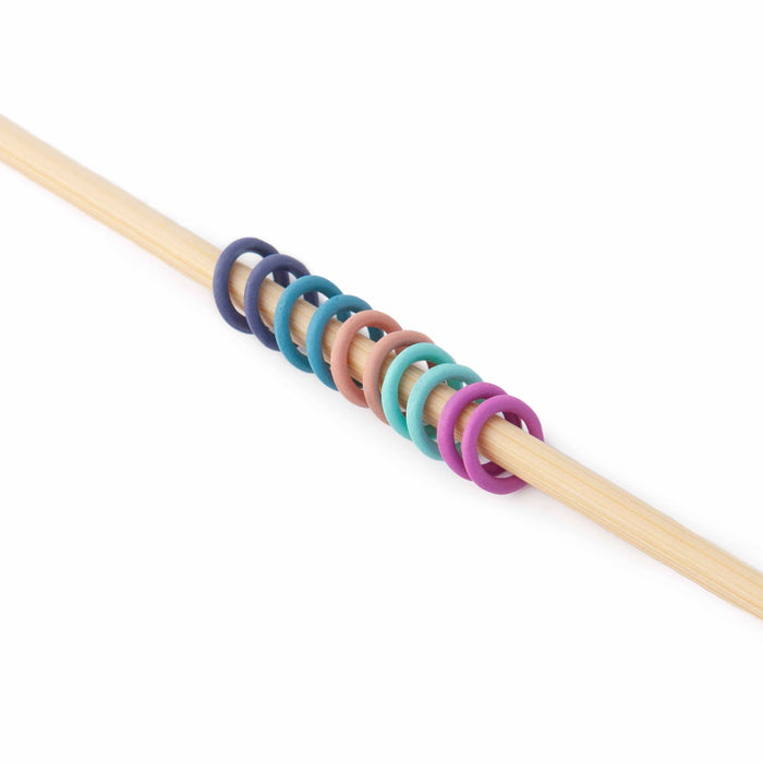 Rainbow-colored ring stitch markers threaded onto a wooden knitting needle on red knit fabric, showing how they sit on the needle during knitting