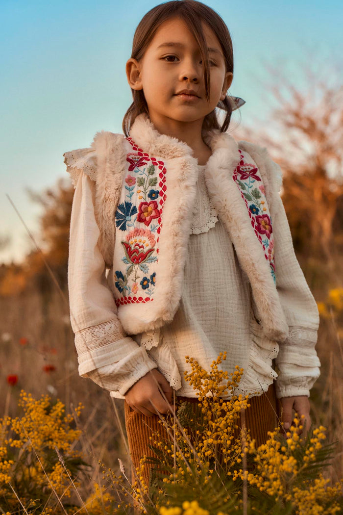 child standing in field wearing faux fur lined vest with floral embroidery in pink, blue, and green.