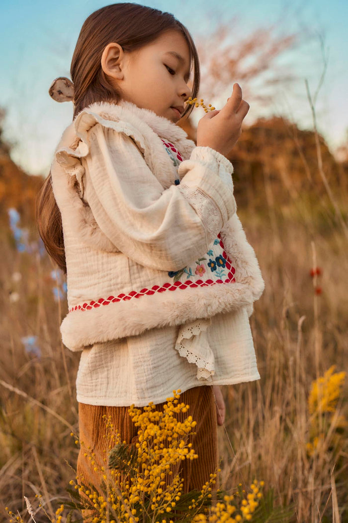 side view of child standing in field wearing faux fur lined vest with floral embroidery in pink, blue, and green.