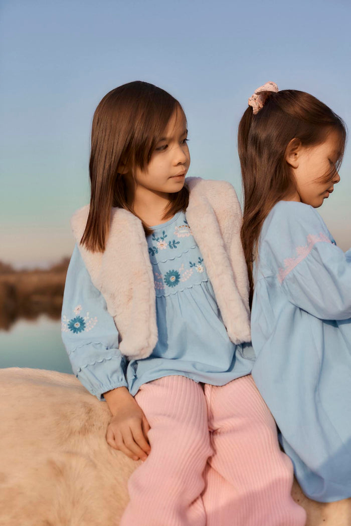 two children sitting on rock outside in front of water. One is wearing a faux fur vest with embroidered denim shirt and pink pants. The other is the side view wearing a denim dress with pink embroidery on sleeve.